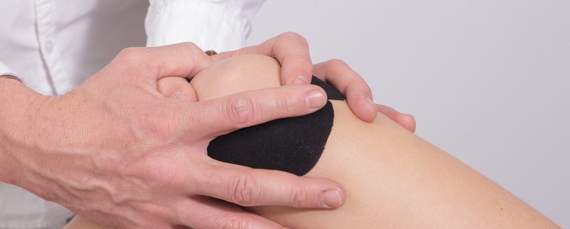 Nahaufnahme einer physiotherapeutischen Behandlung am Knie. Eine Person in einem weißen Kittel hält mit beiden Händen das Knie einer anderen Person, die eine schwarze Bandage oder ein Kinesiotape trägt. Die Szene deutet auf eine Untersuchung oder Therapie zur Schmerzlinderung oder Rehabilitation hin.